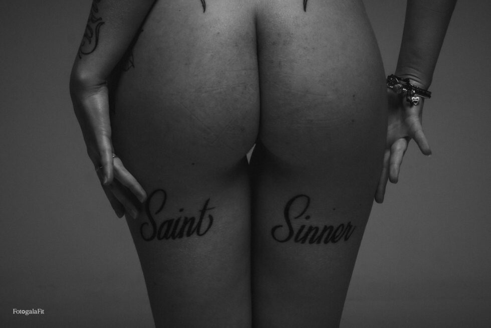 Detalle en blanco y negro de una mujer de espaldas con los tatuajes “Saint” y “Sinner” en la parte posterior de los muslos.