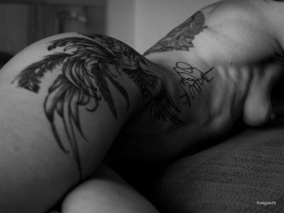 Autorretrato artístico en blanco y negro de un torso recostado con tatuajes de aves. La luz lateral modela el cuerpo y resalta las líneas del hombro y la clavícula, creando una atmósfera íntima y contemplativa.