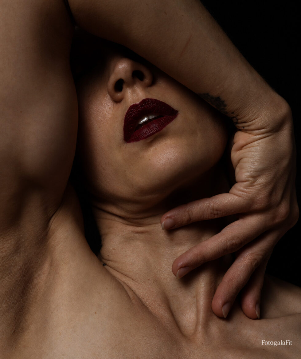 Autorretrato artístico de Fotogalafit con los ojos ocultos tras el brazo y una mano enmarcando el rostro. Luz lateral, fondo negro y labios granates.